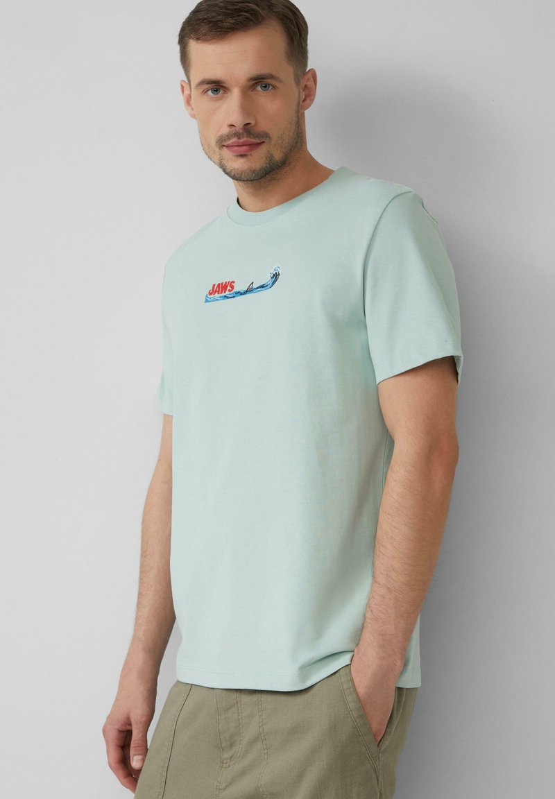 Jasno miętowa koszulka wykonana z bawełny, z małą grafiką "JAWS" w kolorach czerwonym i niebieskim oraz wzorem fal na przodzie, krótki rękaw.