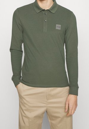 Camisa polo de manga larga en algodón verde oliva, presenta una tapeta de botones, un parche con logo en el pecho y puños ajustados. Se lleva con pantalones beige.