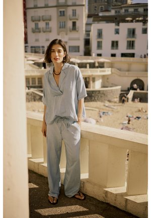 Completo in lino di colore azzurro chiaro, composto da una camicia a maniche corte e pantaloni con cordoncino, entrambi con una vestibilità comoda. Indossato con ciabatte nere.