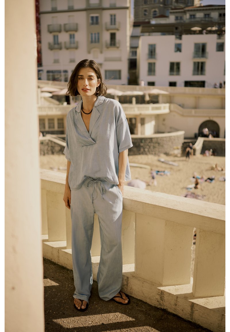 Completo in lino di colore azzurro chiaro, composto da una camicia a maniche corte e pantaloni con cordoncino, entrambi con una vestibilità comoda. Indossato con ciabatte nere.
