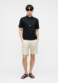 Férfi fekete pólóingben, bézs rövidnadrágban, sötét napszemüvegben, fekete arcmaszkkal a nyakpánton és barna szandálban, kezét a zsebében tartva áll.