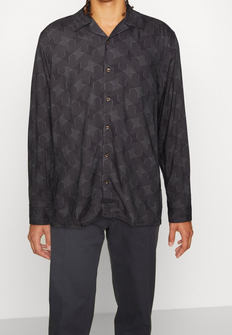Homme portant une chemise noire à manches longues boutonnée avec un motif géométrique en forme de labyrinthe et un pantalon gris foncé sur fond blanc uni.