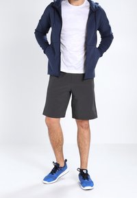 Felpa con zip blu navy, t-shirt bianca, pantaloni corti grigi e sneakers blu. L'outfit presenta un design aderente e materiali sportivi.