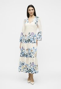 Femme portant une longue robe blanche avec des motifs floraux bleus, violets et beiges, col en V, manches cloches et chaussures blanches pointues, debout.
