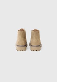 Botas de tobillo de ante color tostado con suela de goma gruesa, costuras verticales y tira trasera para ponerlas y quitarlas con facilidad. Textura suave con acabado mate.