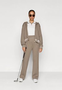 Conjunto de fato de treino castanho claro em malha, com capuz e fecho de correr, e calças de perna larga. Detalhes brancos nos punhos e gola. Modelo segurando um taco de golfe.