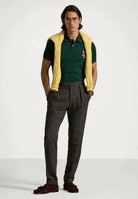 Νέος άνδρας στέκεται με τα χέρια στις τσέπες, φορώντας γυαλιά, πράσινο πόλο μπλουζάκι, γκρι παντελόνι, καφέ λόφερ και ένα κίτρινο πουλόβερ ριγμένο στους ώμους.