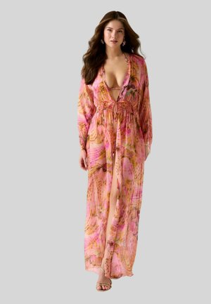 Kobieta w długiej, zwiewnej sukience w różowo-pomarańczowy wzór z głębokim dekoltem i rozcięciem, stojąca na tle szarego tła.