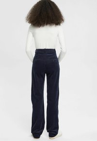 Granatowe sztruksowe spodnie z wysokim stanem i szerokimi nogawkami, z tylnymi kieszeniami i teksturowaną powierzchnią. W tle biała bluzka z długim rękawem.