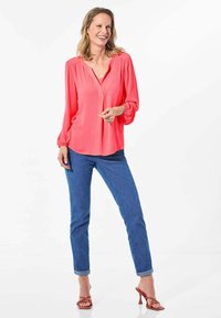 Korallenfarbene Bluse mit langen Ärmeln, lässigem Schnitt und V-Ausschnitt, kombiniert mit blauen aufgerollten Jeans und roten Riemchen-High Heels vor einem weißen Hintergrund.
