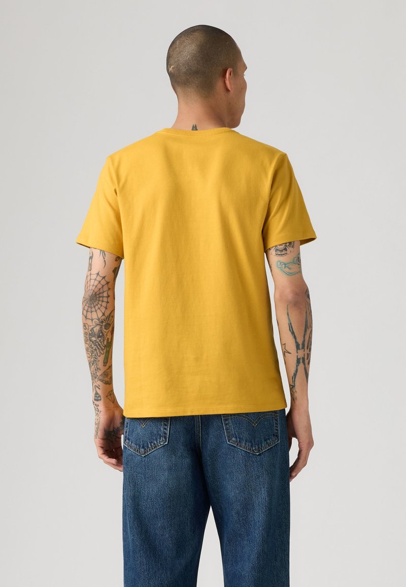 Homme avec des tatouages sur les bras portant un t-shirt jaune moutarde à manches courtes et un jean bleu, vu de dos sur un fond clair uni.