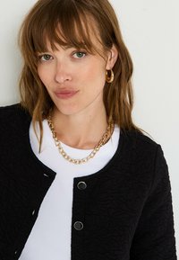 Cárdigan negro texturizado con botones circulares negros, superpuesto sobre una blusa blanca. Presenta un collar de cadena dorada y aretes de aro.