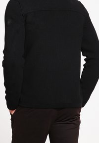 Schwarzer, gerippter Strickpullover mit einem strukturierten Schulterdesign, enganliegenden Ärmeln und einem abgerundeten Saum. Die Rückansicht zeigt ein dezentes Logodetail in der Nähe der Schulter.