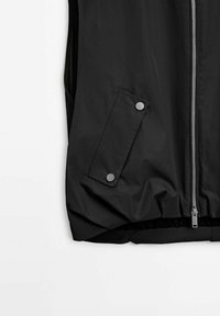 Veste noire sans manches avec fermeture éclair à l'avant et poche latérale sécurisée par deux boutons-pression sur fond blanc.