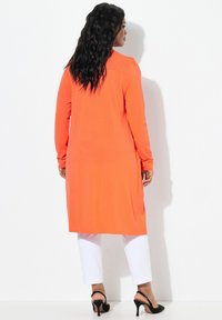 Långärmad orange tunika i mjukt tyg, med sidofickor för ökad rörelsefrihet. Kombinerad med vita byxor och svarta klacksandaler.