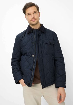 Homme portant une veste matelassée bleu marine sur une chemise marron avec un pantalon beige, debout avec une main dans la poche sur un fond blanc.