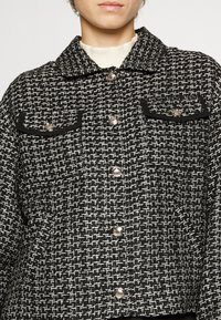 Svart och vit mönstrad jacka av texturerad tyg, med krage, två bröstfickor och silverfärgad knappstängning.