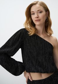 Top noir à une épaule en tissu texturé avec des rayures verticales argentées. La longue manche bouffante et le design court accentuent la silhouette.