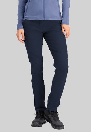 Vrouw draagt marineblauwe slim-fit broek, zwarte wandelschoenen en een lichtblauwe jas met ritssluiting, staand tegen een grijze achtergrond.