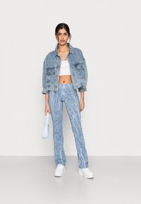 Casaco de ganga, azul claro, de corte oversized, com bolsos frontais. Top branco cortado. Calças azuis com padrão, pernas largas, combinadas com sapatilhas brancas de plataforma e uma pequena mala azul clara.