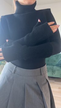 Fekete bordás garbós pulóver hosszú, nyitott ujjakkal, amely egy szürke plisszírozott szoknyával van párosítva. Az anyag puhanak és enyhén texturáltnak tűnik.