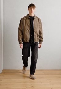 Brun letvægtsjakke over en sort skjorte, parret med sorte jeans og beige sko. Modellen går mod en tekstureret lys baggrund.
