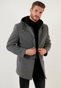 Abrigo de mezcla de lana gris con capucha, que presenta un cierre de botones, dos bolsillos laterales y un forro interior negro. Textura suave, de largo medio.