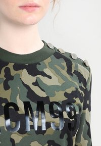 Felpa camouflage in tonalità di verde con dettagli neri, caratterizzata da una stampa lucida "CM99" e bottoni metallici sulle spalle.