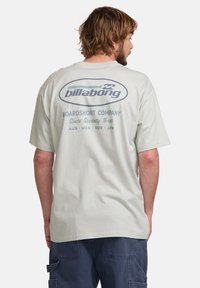 Šviesiai pilkas medvilnės marškinėliai su apvaliu logotipu mėlynos spalvos ant nugaros, kuriame yra tekstas "Billabong" ir "Boardshort Company."