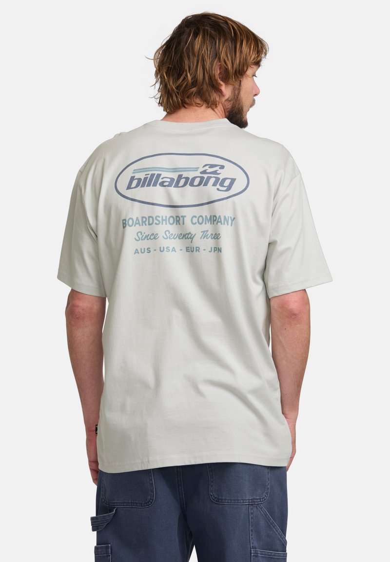 Šviesiai pilkas medvilnės marškinėliai su apvaliu logotipu mėlynos spalvos ant nugaros, kuriame yra tekstas "Billabong" ir "Boardshort Company."
