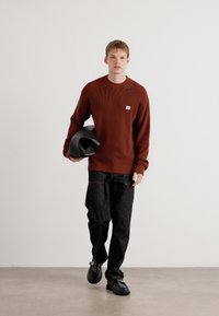 Roestkleurige geribbelde trui, zwarte wijde broek, zwarte schoenen, model met een zwarte tas, minimalistische achtergrond.