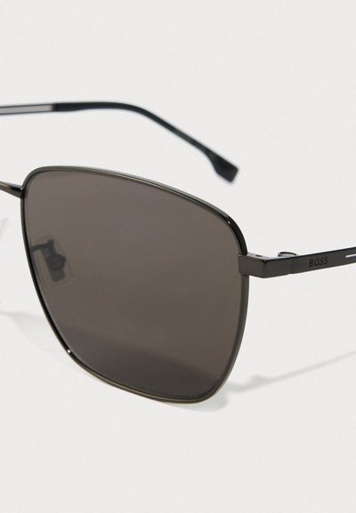 Gros plan de lunettes de soleil à monture noire en métal avec des verres rectangulaires sombres et le logo "BOSS" sur la branche, sur fond blanc.