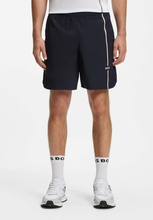 Mann trägt schwarze Sportshorts mit vertikalem weißen Streifen und BOSS-Logo, weiße Socken mit BOSS-Schriftzug und weiße Sneaker.