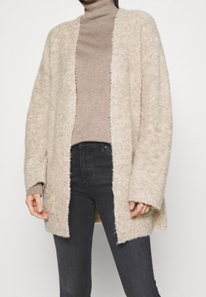 Femme portant un cardigan ouvert beige duveteux sur un pull à col roulé taupe et un jean noir sur un fond uni.