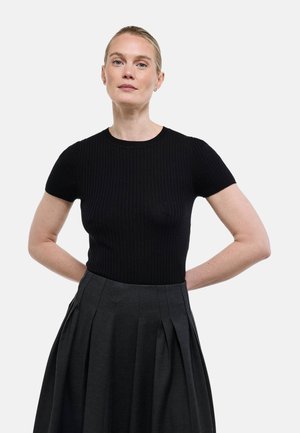 Femme aux cheveux blonds portant un haut noir côtelé à manches courtes et une jupe plissée haute taille foncée, debout avec les mains derrière le dos.