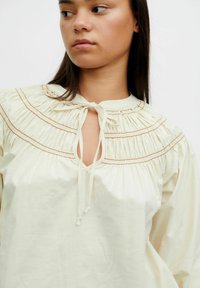 Vrouw in een crèmekleurige blouse met geplooide schouderpartij en strik bij de halsopening, kijkt naar de zijkant tegen een effen witte achtergrond.