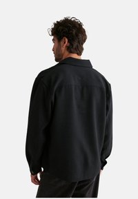 Chaqueta negra texturizada con cuello, mangas largas y un corte relajado, con costuras visibles y puños abotonados.