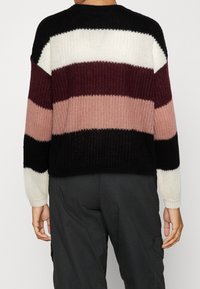 Pull-over en maille présentant des rayures horizontales en noir, blanc, bordeaux et rose poudré. Encolure ronde, coupe décontractée et poignets côtelés.