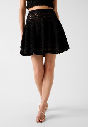 Femme portant une mini-jupe en crochet noire et un crop-top assorti, debout jambes croisées, en sandales dorées à talons hauts à brides sur fond blanc.