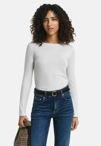 Langsleeve witte katoenen T-shirt gecombineerd met donkerblauwe jeans en een zwarte riem. De outfit wordt aangevuld met een bruine geruite jas.