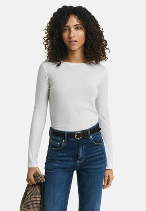 Langärmliges weißes Baumwoll-T-Shirt, kombiniert mit dunkelblauen Jeans und einem schwarzen Gürtel. Das Outfit wird durch eine braune karierten Jacke ergänzt.