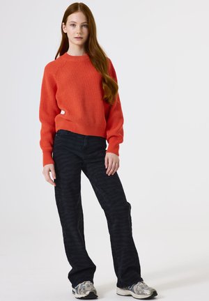Junge Person mit langen roten Haaren, die einen orangefarbenen Strickpullover, schwarze gemusterte Hosen und silberne Turnschuhe trägt und vor einem weißen Hintergrund steht.