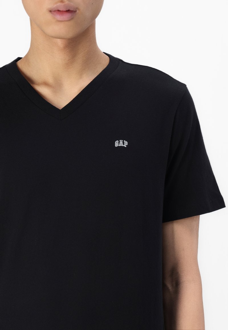 Schwarzes Baumwoll-V-Neck-T-Shirt mit kurzen Ärmeln und einem kleinen weißen "GAP"-Logo auf der linken Brust. Glatte Textur, einfaches Design.