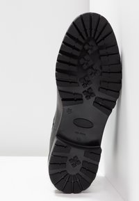 Semelle de botte noire avec un motif profond et rugueux appuyée contre un mur blanc sur un sol blanc.
