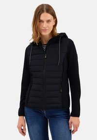 Svart jacka med huva; har en kviltad frampanel, slankt design och dragkedjestängningar; bärs över en randig topp med jeans.