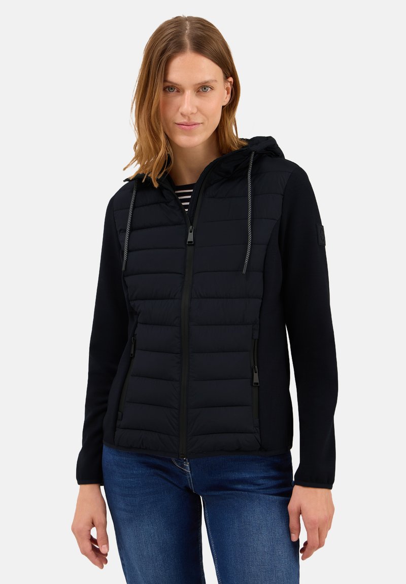 Veste noire avec capuche ; présente un panneau avant matelassé, un design épuré et des fermetures éclairs ; portée sur un haut rayé avec un jean.