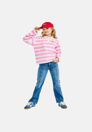 Młoda dziewczyna w czerwonej czapce, różowo-białej pasiastiej bluzce z długim rękawem, niebieskich dżinsach i niebieskich tenisówkach, pozująca z jedną ręką na biodrze.