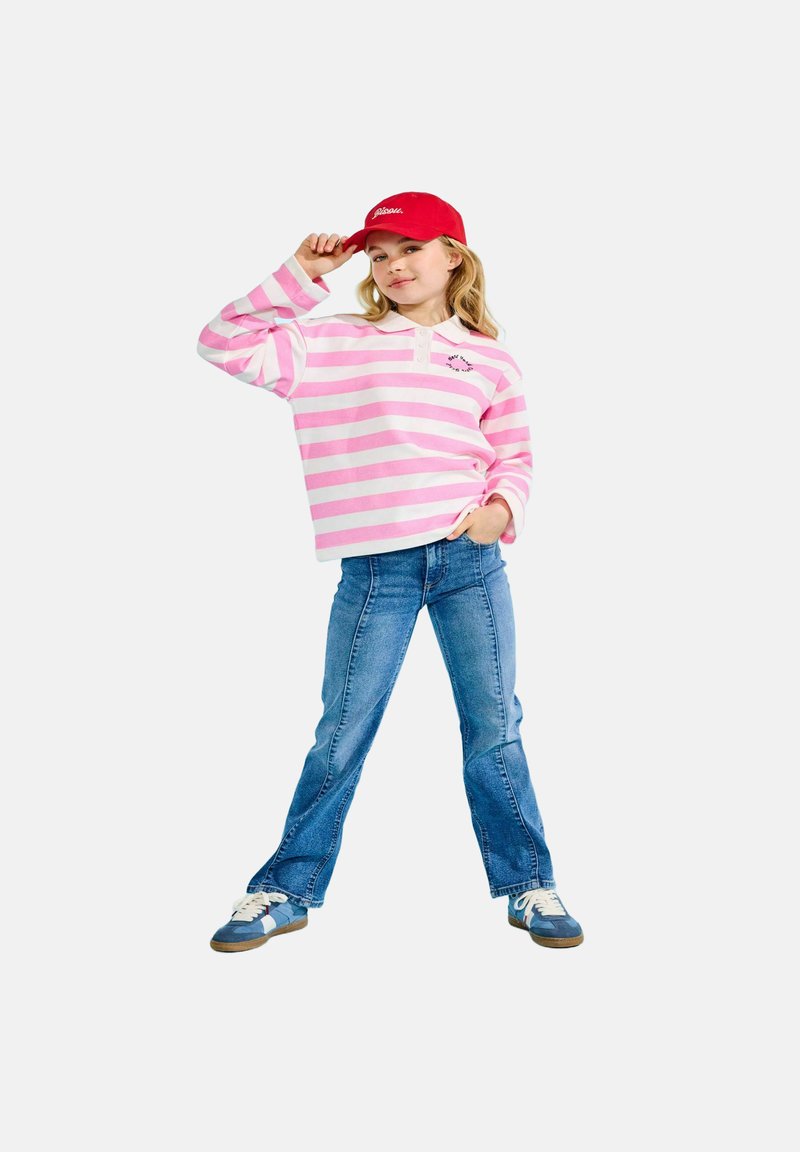 Jeune fille portant une casquette rouge, un t-shirt à manches longues rose et blanc à rayures, un jean bleu et des baskets bleues, posant avec une main sur la hanche.