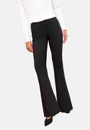 Frau trägt schwarze Schlaghose, weiße Bluse mit langen Ärmeln und schwarze spitze High Heels, steht vor einem einfarbigen Hintergrund.