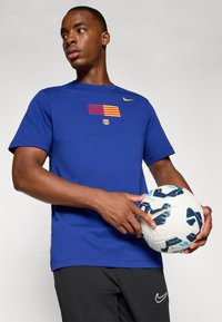 Blå bomulds t-shirt med et horisontalt stribet Barcelona-logo og guld Nike swoosh. Modellen holder en hvid og blå mønstret fodbold.